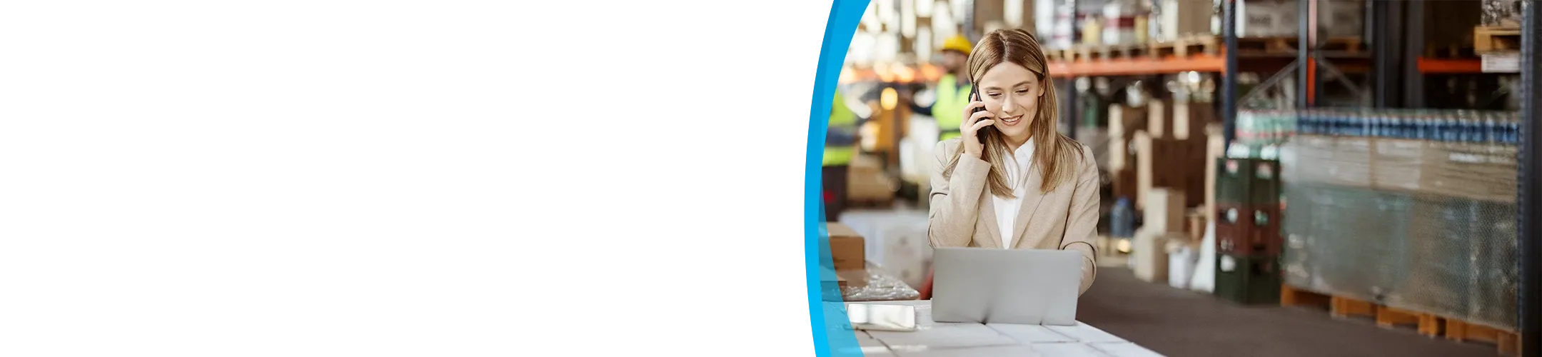 A smiling female manager in a business suit is talking on a phone and working on a laptop on top of wrapped pallets in a busy warehouse.